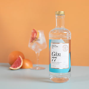 Bottle of Gin 77 in the fore ground, with a glass of gin and tonic in the back ground. Garnished of pink grapefruit and some pink grapefruit sat on the side. 